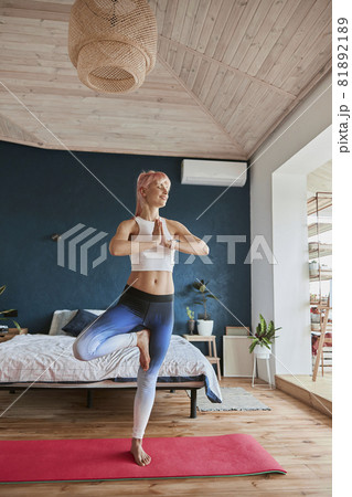 Joyful yoga practitioner in comfortable sportswear stands in tree position in light room Joyful yoga practitioner in comfortable sportswear stands in tree position in light room 81892189
