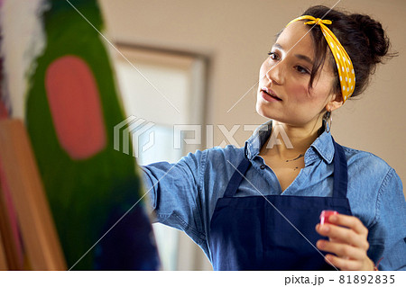 Portrait of young woman in apron looking inspired while working on abstract painting at cozy workshop 81892835