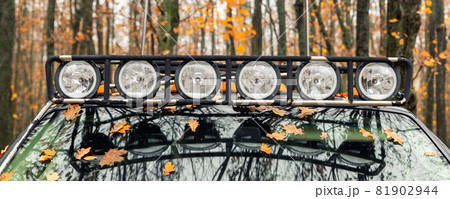Close-up detail view of custom made roof rack bar with extra headlight mounted on roof of heavy duty pick up suv car against foggy autumn forest. Fallen leaves vehicle windshield. Fall weather drive Close-up detail view of custom made roof rack bar with extra headlight mounted on roof of heavy duty pick up suv car against foggy autumn forest. Fallen leaves vehicle windshield. Fall weather drive 81902944