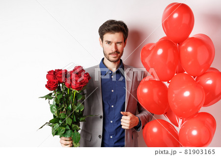 Confident boyfriend in suit going on romantic date, winking at camera, holding bouquet of red roses, standing near heart balloon and winking, standing over white background 81903165