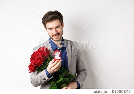 Romantic man with boquet of red roses asking to marry him, holding engagement ring and looking confident at camera, standing in suit over white background 81906879