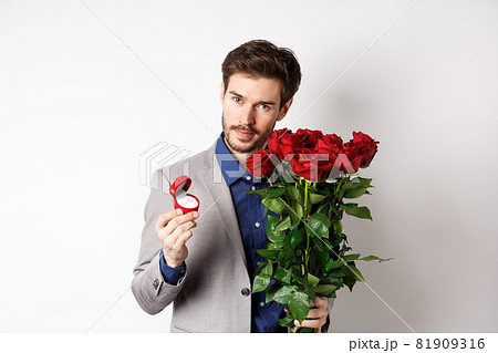 Handsome man in suit, showing engagement ring and looking romantic at camera, standing with red roses over white background. Valentines day and love concept Handsome man in suit, showing engagement ring and looking romantic at camera, standing with red roses over white background. Valentines day and love concept 81909316