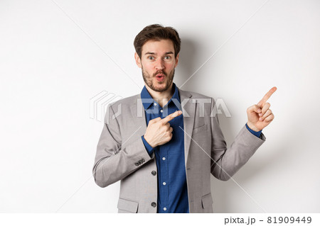 Excited handsome caucasian businessman in suit pointing right at logo, say wow and look amazed, standing on white background 81909449