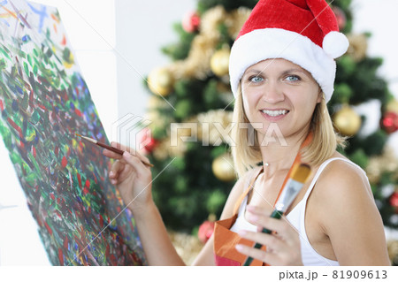 Smiling woman artist in santa claus hat draws picture against background of Christmas tree 81909613