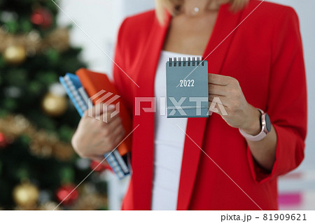 Businesswoman in red jacket holds calendar for 2022 against background of New Year tree Businesswoman in red jacket holds calendar for 2022 against background of New Year tree 81909621