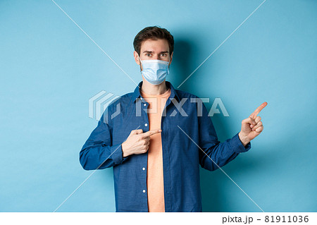 Covid and healthcare concept. Handsome modern guy in medical mask pointing at empty space aside, standing on blue background 81911036