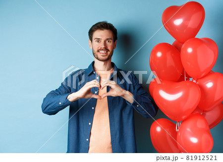 Happy valentines day. Boyfriend in love showing heart gesture to lover and smiling, standing near red balloons on blue background Happy valentines day. Boyfriend in love showing heart gesture to lover and smiling, standing near red balloons on blue background 81912521