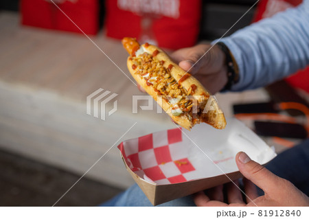 hand holds fried sausage in dough with ketchup in paper red white packaging on the street. hand holds fried sausage in dough with ketchup in paper red white packaging on the street. 81912840