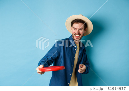 Happy man laughing and playing frisbee, throwing at friend and smiling, standing in summer hat on blue background Happy man laughing and playing frisbee, throwing at friend and smiling, standing in summer hat on blue background 81914937