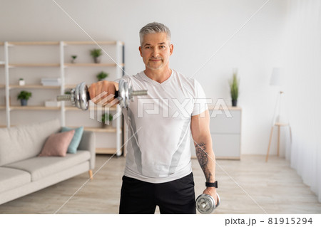 Domestic training with weights. Positive mature man doing exercises with dumbbells, strengthening his body at home 81915294
