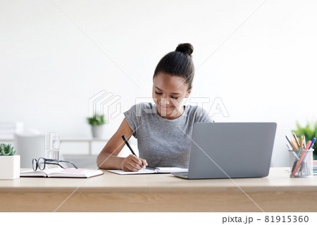 Smiling young african american girl pupil writes in notebook at computer, studies Smiling young african american girl pupil writes in notebook at computer, studies 81915360