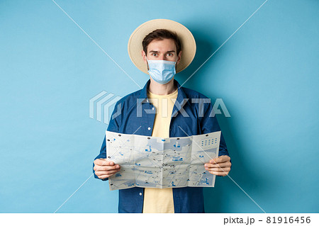 Covid-19, pandemic and travel concept. Happy tourist on summer vacation wearing medical mask and straw hat, holding road map, standing on blue background 81916456