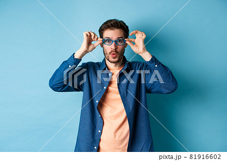 Funny young man trying new sunglasses, looking surprised at camera, standing on blue background Funny young man trying new sunglasses, looking surprised at camera, standing on blue background 81916602