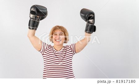 An elderly woman in fighting gloves raises her hands up on a white background. Victory in a duel An elderly woman in fighting gloves raises her hands up on a white background. Victory in a duel 81917180