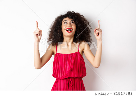 Excited caucasian woman with curly hair, red lips, looking and pointing up with amazed smile, checking out promo offer, white background Excited caucasian woman with curly hair, red lips, looking and pointing up with amazed smile, checking out promo offer, white background 81917593