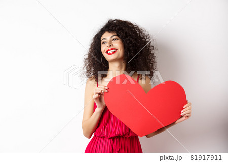 Romantic woman in dress and makeup, looking aside with dreamy face and love, showing big red valentines heart card, standing on white background 81917911