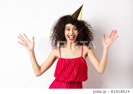 Cheerful young woman in red dress, celebrating birthday, wearing party hat and smiling, screaming of joy, standing on white background Cheerful young woman in red dress, celebrating birthday, wearing party hat and smiling, screaming of joy, standing on white background 81918624