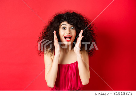 Close-up of surprised beautiful woman, screaming of amazement and looking at promo offer, standing in red dress and lipstick, studio background Close-up of surprised beautiful woman, screaming of amazement and looking at promo offer, standing in red dress and lipstick, studio background 81918875