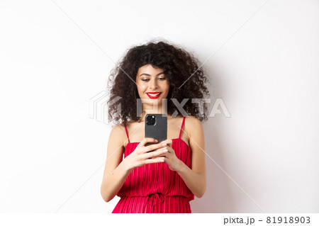 Happy fashionable woman taking photo on smartphone, photographing something on mobile and smiling, standing in red dress on white background 81918903