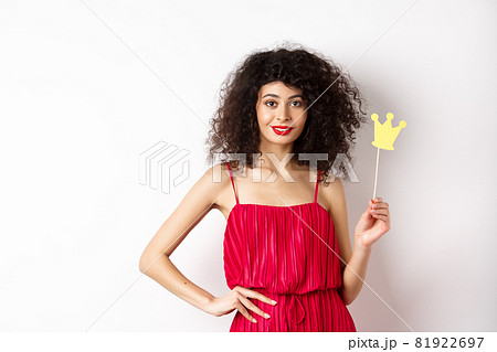 Stylish young woman in red dress, feeling confident and sassy, holding crown and smiling, standing over white background Stylish young woman in red dress, feeling confident and sassy, holding crown and smiling, standing over white background 81922697