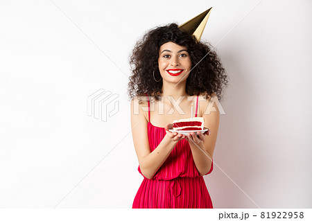 Beautiful woman in red dress, wearing party hat and celebrating birthday, holding b-day cake and making wish, smiling at camera, standing on white background Beautiful woman in red dress, wearing party hat and celebrating birthday, holding b-day cake and making wish, smiling at camera, standing on white background 81922958