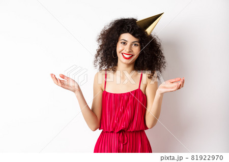 Beautiful caucasian woman in red dress, celebrating holiday, wearing party hat and smiling, standing on white background Beautiful caucasian woman in red dress, celebrating holiday, wearing party hat and smiling, standing on white background 81922970