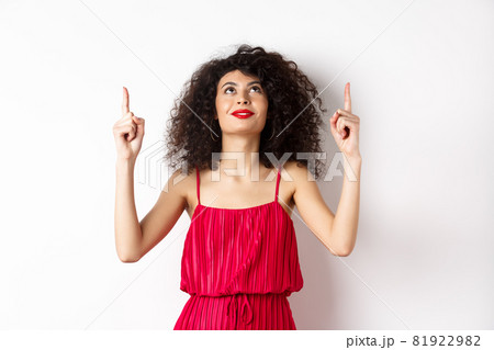 Dreamy young woman in trendy red dress, looking up and pointing at logo, smiling happy, standing over white background Dreamy young woman in trendy red dress, looking up and pointing at logo, smiling happy, standing over white background 81922982