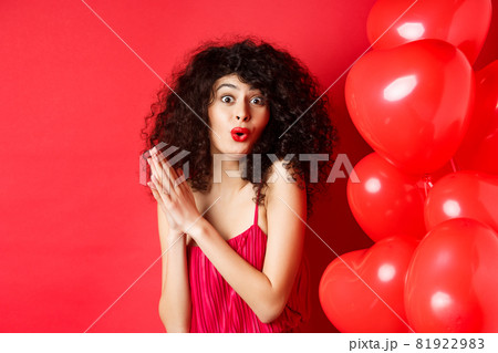 Excited beautiful woman with curly hair, standing near heart balloons and rubbing palms together, expect good deal or relish, red background Excited beautiful woman with curly hair, standing near heart balloons and rubbing palms together, expect good deal or relish, red background 81922983