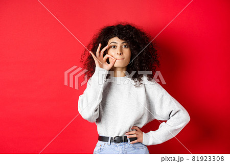 Image of cute girl with curly hair making promise to stay quiet, zipping lips, making seal, hiding a secret, standing dumb on red background Image of cute girl with curly hair making promise to stay quiet, zipping lips, making seal, hiding a secret, standing dumb on red background 81923308