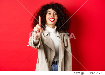 Cheerful woman in trench coat, showing number one finger and smiling, standing on red background Cheerful woman in trench coat, showing number one finger and smiling, standing on red background 81923473