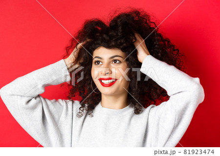 Beauty. Close-up portrait of carefree woman touching her curly hair and looking happy aside at logo, smiling with white teeth and red lips, studio background Beauty. Close-up portrait of carefree woman touching her curly hair and looking happy aside at logo, smiling with white teeth and red lips, studio background 81923474