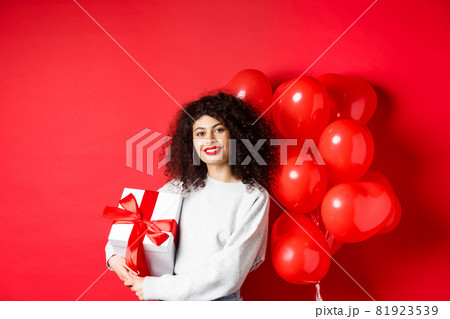 Beautiful caucasian woman with dark curly hair and red lips, standing with romantic hearts balloons and present in box, receive gift from lover, standing on studio background 81923539
