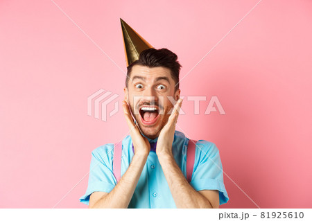 Holidays and celebration concept. Close-up of funny guy in birthday hat shouting surprised, screaming with joy, receiving awesome gift, standing happy on pink background 81925610
