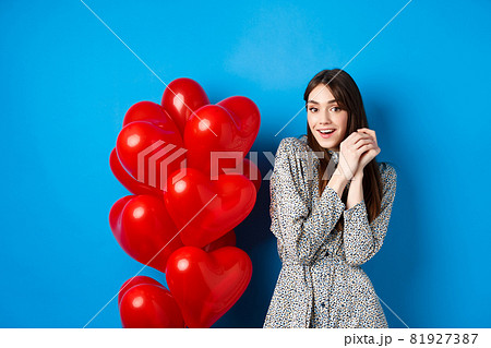 Valentines day. Lovely girlfriend ind ress smiling happy at camera, celebrating lovers holiday, standing near heart balloons, blue background Valentines day. Lovely girlfriend ind ress smiling happy at camera, celebrating lovers holiday, standing near heart balloons, blue background 81927387