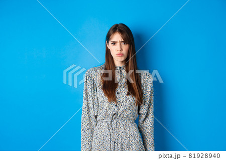 Moody frowning girl in dress looking at unfair thing, standing offended or upset against blue background 81928940