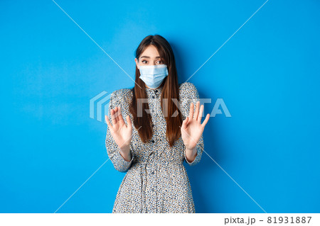 Covid-19, pandemic lifestyle concept. No thank you. Reluctant and worried girl in medical mask asking non-masker to stay away, showing block gesture, tell stop, blue background 81931887