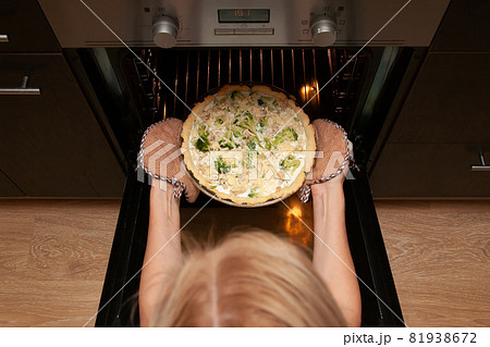 Woman takes an open shortbread pie with chicken and broccoli, sprinkled with cheese out of the oven in her kitchen. 81938672