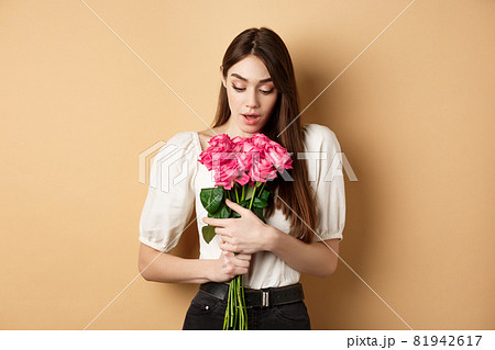Valentines day. Surprised tender girl looking at beautiful pink roses, receive romantic gift from lover, standing on beige background Valentines day. Surprised tender girl looking at beautiful pink roses, receive romantic gift from lover, standing on beige background 81942617