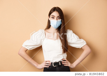 Covid-19 and lifestyle concept. Woman in medical mask looking alarmed at camera, standing on beige background 81943802