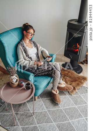 woman by the fireplace, sitting in a cozy armchair, with a warm woman by the fireplace, sitting in a cozy armchair, with a warm 81944453