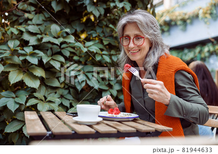 Smiling mature female guest with glasses eats toast at table on outdoors cafe terrace Smiling mature female guest with glasses eats toast at table on outdoors cafe terrace 81944633