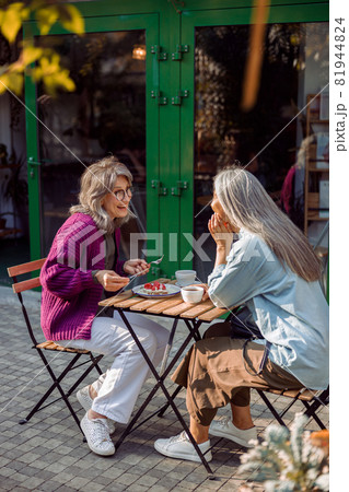 Joyful mature woman with friend small table with dessert and drinks outdoors 81944824