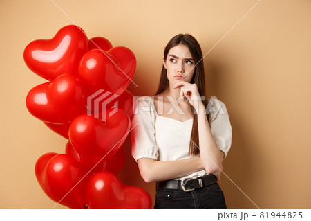 Valentines day and love concept. Thoughtful young woman standing near hearts balloons and looking aside with doubtful face, having assumptions, beige background Valentines day and love concept. Thoughtful young woman standing near hearts balloons and looking aside with doubtful face, having assumptions, beige background 81944825