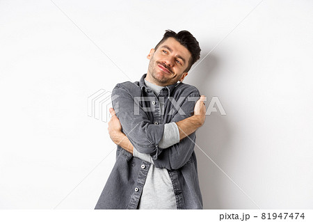 Happy romantic guy hugging himself with closed eyes and smile, loving own body, daydreaming about lover, standing on white background 81947474