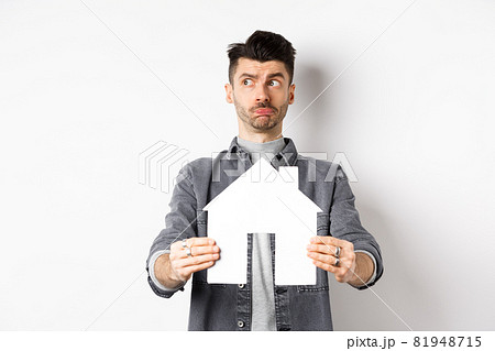 Real estate and insurance concept. Indecisive guy look aside pensive and showing paper house cutout, thinking of buying property, standing against white background Real estate and insurance concept. Indecisive guy look aside pensive and showing paper house cutout, thinking of buying property, standing against white background 81948715