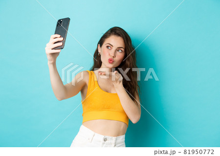 Technology and lifestyle concept. Stylish beautiful woman posing for selfie, sending air kiss at camera, using photo filters app on mobile phone, standing against blue background Technology and lifestyle concept. Stylish beautiful woman posing for selfie, sending air kiss at camera, using photo filters app on mobile phone, standing against blue background 81950772