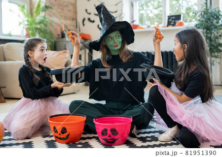 Young female in witch attire putting treats in hats of two adorable girls Young female in witch attire putting treats in hats of two adorable girls 81951390