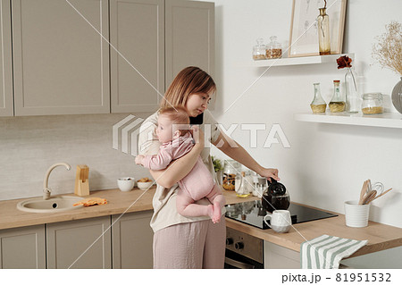 Young woman with baby pouring tea into mug and talking on smartphone 81951532