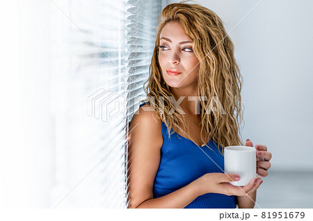 Young woman leans back to the window and and drink some beverage while she is looking to a point. Young woman leans back to the window and and drink some beverage while she is looking to a point. 81951679