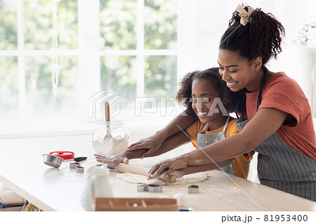 Cheerful black mother helping daughter rolling pasrty dough 81953400